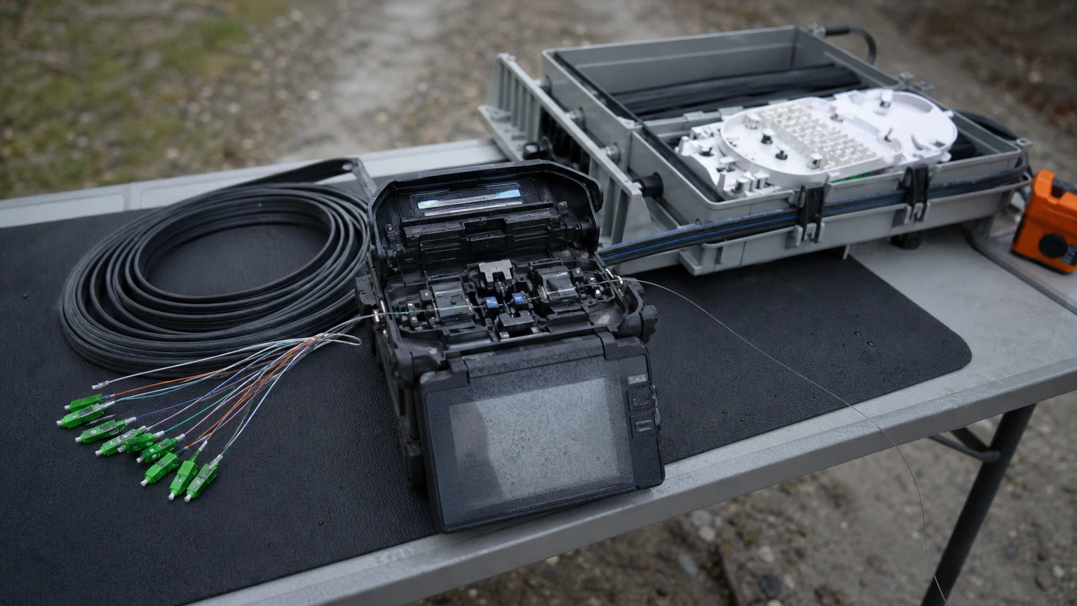 A fiber optic fusion splicer with two bare fibers aligned in the v-groove beside a coil of drop cable and an open splice closure